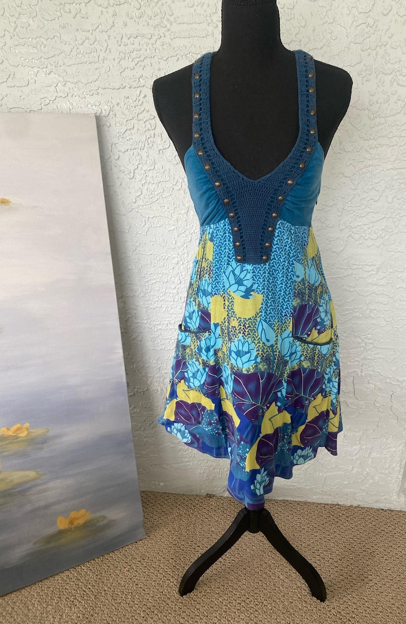 May include: A blue and yellow floral print dress with a halter top. The top is a dark blue crochet with bronze-colored studs. The dress has a V-neck and two small pockets. The dress is displayed on a black mannequin.