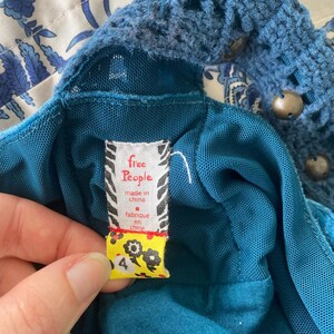以下が含まれることがあります： かぎ針編みのネックラインと「free People, made in china, fabrique en chine」と書かれたタグが付いた、ティール色のドレスのクローズアップ。タグは黄色で、花の模様と数字の4が付いています。