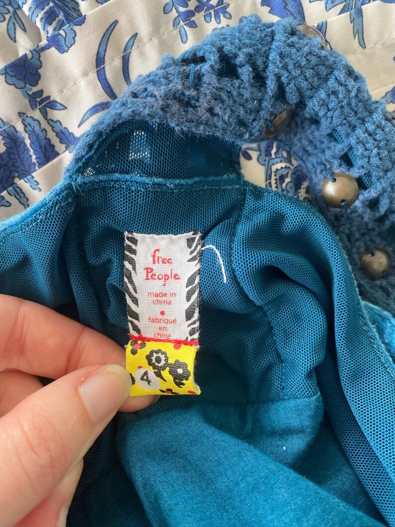 以下が含まれることがあります： かぎ針編みのネックラインと「free People, made in china, fabrique en chine」と書かれたタグが付いた、ティール色のドレスのクローズアップ。タグは黄色で、花の模様と数字の4が付いています。