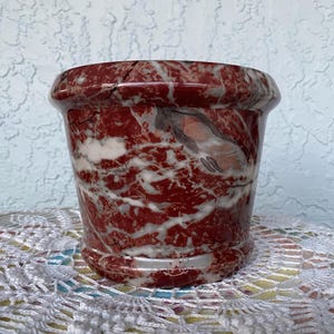 May include: A decorative, cylindrical flower pot with a flared rim. The pot features a striking pattern of red and white marbling. The pot is made of a solid material and has a smooth, polished finish. The pot is suitable for indoor or outdoor use.