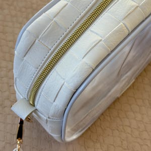 May include: A white rectangular cosmetic bag with a woven leather design. The bag features a gold zipper and a small strap with gold hardware. The clear side panel allows visibility of the contents.