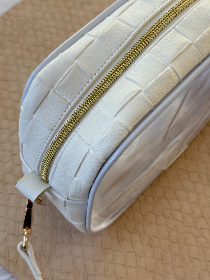 May include: A white rectangular cosmetic bag with a woven leather design. The bag features a gold zipper and a small strap with gold hardware. The clear side panel allows visibility of the contents.
