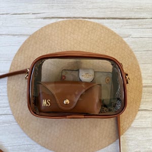 May include: A clear crossbody bag with brown leather trim and a brown strap. Inside the bag are a brown sunglasses case with "MS" in gold lettering, and a floral wallet. The bag is on a round, tan surface.