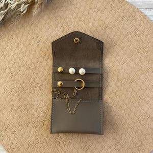 May include: A small, open, gray leather jewelry organizer with gold-colored studs, a gold hoop earring, a gold chain necklace, and pearl earrings. The organizer is open, revealing the jewelry inside. The background is a beige, textured surface.