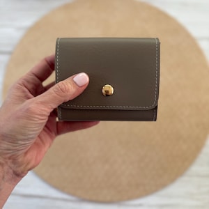 May include: A small, square, taupe-colored wallet with a gold-colored snap closure. The wallet has white stitching around the edges. The wallet is held in a person's hand.
