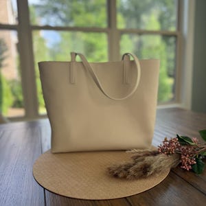 May include: A beige tote bag with two handles sits on a round, textured placemat. The bag is on a wooden table, with a decorative floral arrangement to the right. A window with a view of trees is in the background.