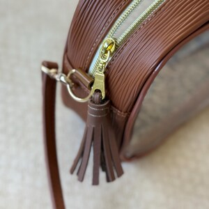 May include: A brown, round handbag with a textured exterior and a gold zipper. The bag features a small, brown tassel and a matching shoulder strap. The interior is clear, allowing visibility of the contents.