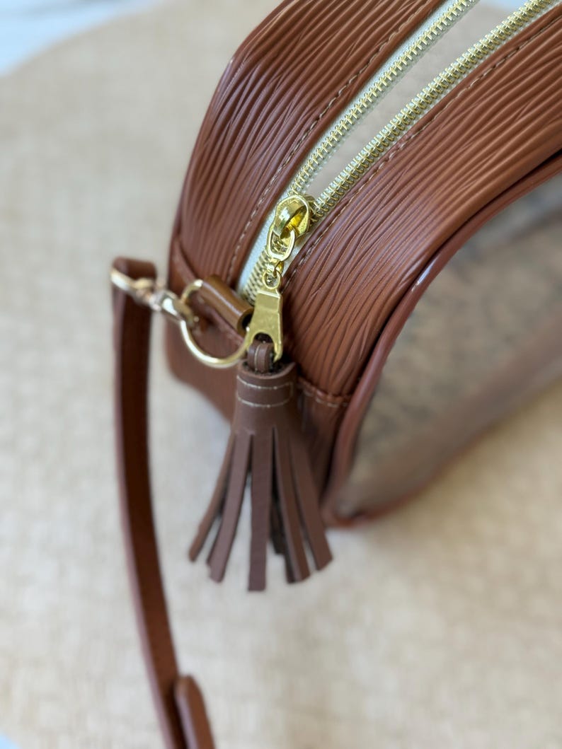 May include: A brown, round handbag with a textured exterior and a gold zipper. The bag features a small, brown tassel and a matching shoulder strap. The interior is clear, allowing visibility of the contents.