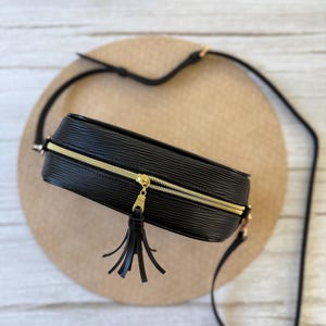 May include: Black crossbody bag with a gold zipper and tassel. The bag has a textured surface and a long, adjustable strap. The bag is rectangular and appears to be made of leather or a similar material. The zipper is gold-colored.