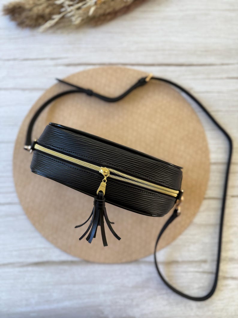 May include: Black crossbody bag with a gold zipper and tassel. The bag has a textured surface and a long, adjustable strap. The bag is rectangular and appears to be made of leather or a similar material. The zipper is gold-colored.