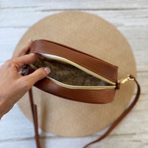 May include: A brown, textured crossbody bag with a gold zipper and strap. The bag is open, revealing a patterned interior. The bag is sitting on a beige surface.