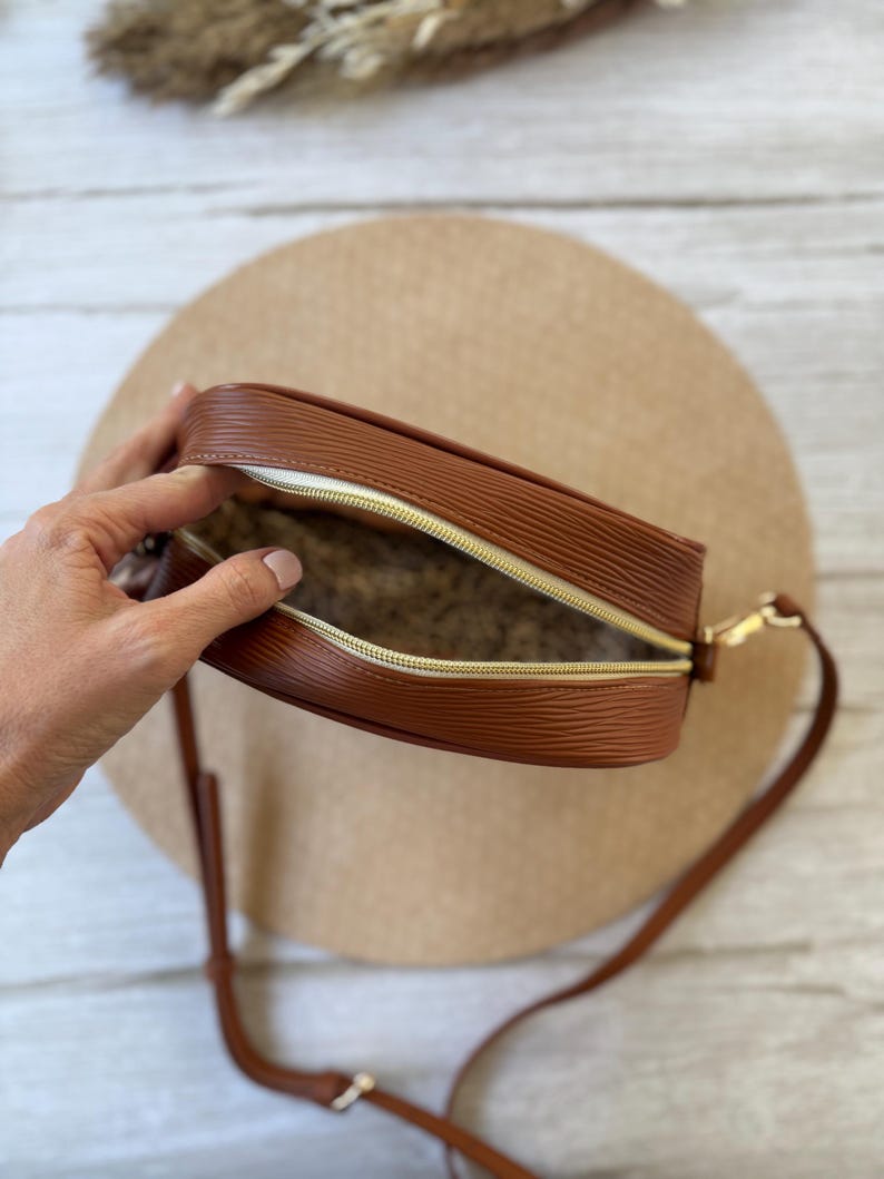 May include: A brown, textured crossbody bag with a gold zipper and strap. The bag is open, revealing a patterned interior. The bag is sitting on a beige surface.