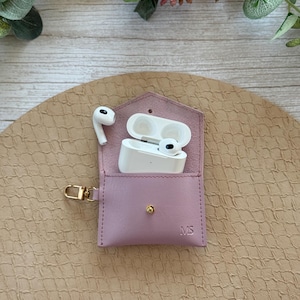 May include: A light purple leather AirPod case with a gold-colored clasp. The case is open, revealing white AirPods and their charging case. The initials "MS" are embossed on the front. The case is on a textured, tan surface.