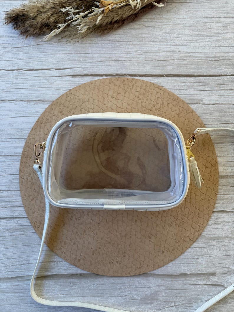 May include: A white, quilted crossbody bag with a clear front panel. The bag has a white strap, gold-colored hardware, and a small tassel. The bag is rectangular and sits on a round, textured, tan surface.