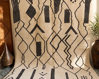 May include: A large, rectangular, cream-colored area rug featuring a bold, black geometric pattern of jagged lines, diamonds, and solid rectangular blocks. The rug has a thick, textured pile and is finished with a knotted fringe along the bottom edge.