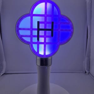 May include: A white and silver light-up wand with a four-leaf clover-shaped head. The head features a grid pattern and a central "H" design, illuminated with blue and white light. The wand has a white handle and a wrist strap.