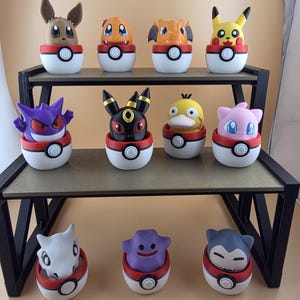 May include: A collection of ten colorful Pokémon character figures displayed on a two-tiered black metal shelf. Each figure is set inside a white and red Poké Ball-themed base. The figures include Pikachu, Eevee, and other popular characters.