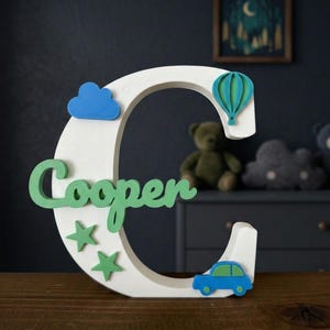 May include: A white wooden letter "C" with the name "Cooper" in green script. The letter is decorated with a blue cloud, green stars, a blue and green hot air balloon, and a blue car. The decor is on a wooden surface.