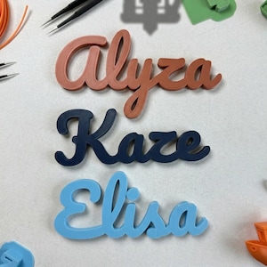 May include: Three custom name signs in coral, navy blue, and light blue script. The names are Alyza, Kaze, and Elisa. The image also includes orange filament, tweezers, and small colorful plastic pieces, suggesting a 3D printing theme.