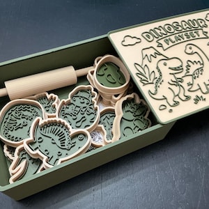 May include: A green rectangular storage box containing various dinosaur-shaped cookie cutters and a small wooden rolling pin. The lid is light beige with dark green embossed illustrations of dinosaurs and the text DINOSAUR PLAYSET.