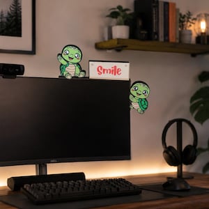 May include: A computer monitor with a webcam, keyboard, and headphones on a wooden desk. Two cartoon turtle stickers are on the monitor. A small sign with the word "Smile" is on the monitor. A small potted plant is on the desk.