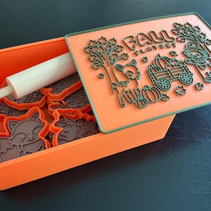 May include: An orange storage box containing brown cookie cutters and a white wooden rolling pin. The lid is orange with a green raised design featuring a deer, a wagon with pumpkins, a scarecrow, trees, and the text FALL PLAYSET.