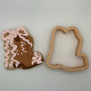May include: A decorated cowboy boot cookie with pink icing and a matching cookie cutter. The boot is brown with white accents and a pink bow. The cookie cutter is a light brown color.