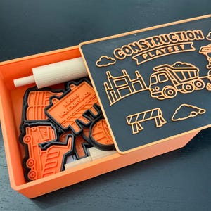 May include: An orange rectangular storage box containing several orange and black construction-themed cookie cutters and a white rolling pin. The lid is black with orange outlines of a dump truck, a crane, a worker, and the text CONSTRUCTION PLAYSET.
