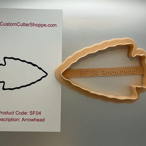 May include: An arrowhead-shaped cookie cutter in a light tan color. The cutter has a handle with the text "SF.04 Arrowhead". A white card displays a black outline of the arrowhead and the text "CustomCutterShoppe.com".