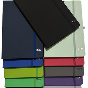 May include: A selection of coloured notebooks, including black, navy, and green, with elastic closures. Each notebook features initials or a name on the cover. The notebooks are stacked, with a black notebook on top. Measurements are not visible.
