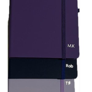 May include: A selection of personalised notebooks in shades of purple, navy, grey, pale green, and blue. Each notebook features a coordinating elastic closure and a name or initials printed on the cover, including "M.K" and "Sophie."