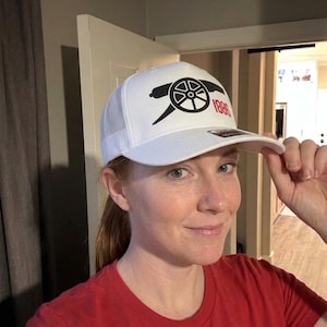 May include: A white trucker-style baseball cap featuring a black cannon graphic and the red text 1886 on the front panel. The cap has a structured front, a curved brim, and a mesh back panel for ventilation, with a small sticker on the brim.