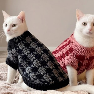 May include: Two white cats wearing crocheted jumpers. One cat wears a black and grey chequered jumper, while the other cat wears a pink and dark red chequered jumper. Both jumpers feature short sleeves and a textured, woven yarn construction.