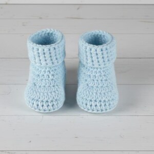 May include: A pair of light blue crochet baby booties. The booties have a rolled-down cuff and a textured stitch pattern. They are made of soft yarn and are designed for infants. The booties are displayed on a white wooden surface.