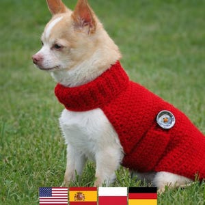 May include: A small dog wearing a bright red knitted sweater with a high collar. The sweater has a decorative button on the side. The dog is sitting on green grass.