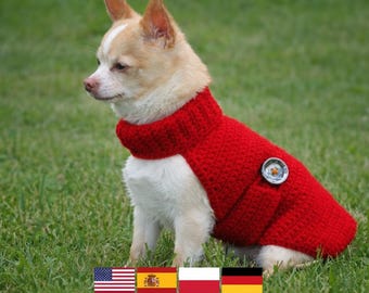 May include: A small dog wearing a bright red knitted sweater with a high collar. The sweater has a decorative button on the side. The dog is sitting on green grass.