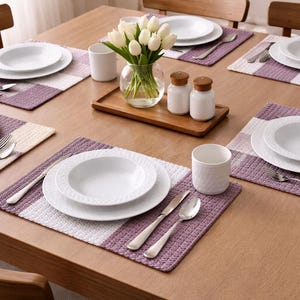 Puede incluir: Una mesa de comedor de madera para seis personas con platos blancos, cubiertos y tazas blancas. Cada cubierto descansa sobre un mantel individual a rayas moradas y blancas. Un jarrón de tulipanes blancos y saleros y pimenteros están en el centro.