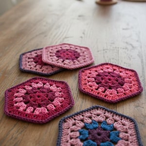 May include: A collection of handmade, hexagonal crochet coasters in shades of pink, purple, and blue. The coasters are arranged on a wooden table, showcasing their intricate floral-like patterns. A mug of tea sits in the background.