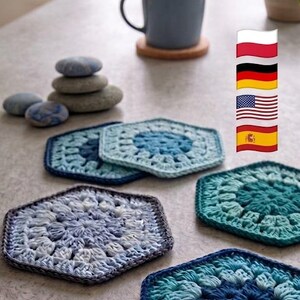 May include: A collection of four hexagonal crochet coasters in various shades of blue and teal, featuring intricate yarn patterns. The coasters are arranged on a surface alongside a ball of beige yarn, a grey mug on a wooden coaster, and smooth stones.