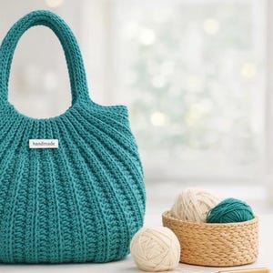 Puede incluir: Bolso de mano de ganchillo hecho a mano, de color verde azulado, con un diseño estructurado y dos asas. Una pequeña etiqueta rectangular dice "handmade". Una pequeña cesta contiene ovillos de lana en crema y verde azulado.