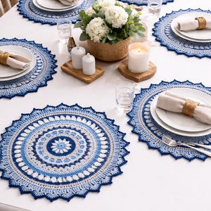 May include: A dining table setting with six white plates, each on a blue and white crocheted placemat. Napkins are rolled and tied with twine. A basket of white flowers, candles, and salt and pepper shakers complete the scene.
