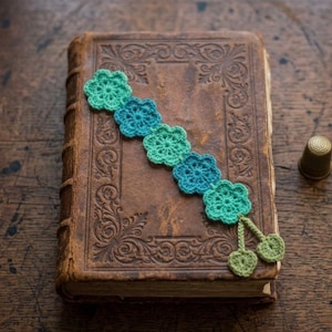 May include: A handmade crochet bookmark in shades of green and blue, shaped like flowers, rests on a vintage book with embossed details. A gold thimble sits nearby on a wooden surface. The bookmark has two green heart-shaped tassels.