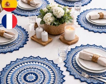 May include: A dining table set with six blue and white crochet placemats. Each placemat has a circular design with a scalloped edge. White plates, napkins, and silverware are arranged on the placemats. A basket of white flowers and a lit candle are in the center.