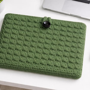 Könnte beinhalten: Eine grüne, gehäkelte Laptoptasche mit strukturiertem Muster und einem schwarzen Knopfverschluss. Die Hülle ist rechteckig und scheint handgefertigt zu sein. Die Laptoptasche liegt auf einer weißen Oberfläche mit einem Laptop, einem Notizbuch und einem Stift im Hintergrund.