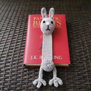 May include: A handmade gray crochet bunny bookmark with black eyes, a pink nose, and long ears. The bookmark is placed in a red book with the title "Fantastic Beasts and Where to Find Them" by J.K. Rowling.