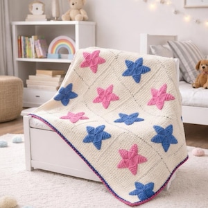 May include: Cream-coloured, hand-crocheted blanket with a repeating pattern of pink and blue stars. The blanket has a dark blue and purple border. It is draped over a white bed frame, creating a cosy and inviting aesthetic. The blanket is approximately 120 cm x 150 cm.