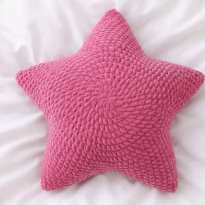 May include: A pink, star-shaped cushion with a textured surface, possibly knitted or crocheted. The cushion is set against a backdrop of white, creased fabric. This decorative cushion would be a charming addition to a bedroom or living space.