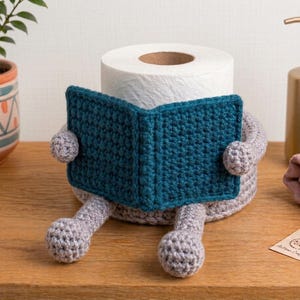 Puede incluir: Un soporte de ganchillo para papel higiénico con una base gris claro, dos piernas y dos brazos que sostienen una cubierta rectangular en forma de libro de color azul cerceta oscuro. El soporte rodea un rollo de papel higiénico blanco.