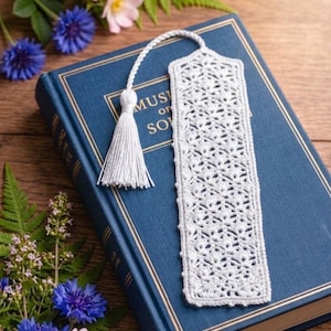 Könnte beinhalten: Handgefertigter, weißer Häkel-Lesezeichen mit Quaste, auf einem blauen Buch. Das Lesezeichen hat ein Lochmuster. Blumen und Blätter umgeben das Buch und bilden eine dekorative Szene.