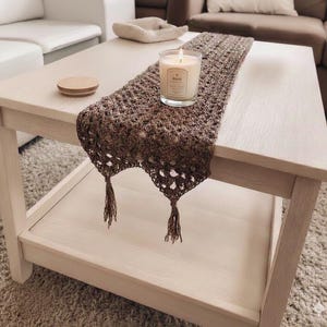 Peut inclure: Une table basse en bois clair avec un chemin de table en crochet marron. Une bougie allumée dans un pot en verre est posée sur le chemin de table. Le chemin de table a des glands aux extrémités. Un couvercle en bois est posé sur la table.
